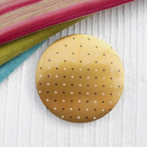 May include: A circular, gold-toned compact featuring a pattern of small black and white stars. The compact sits on a white surface, with colourful fabric in pink, green, and blue in the background.