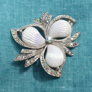 Könnte beinhalten: Eine silberne Brosche mit einem weißen, irisierenden Blumendesign. Die Blume hat drei Blütenblätter und ist von Strasssteinen umgeben.