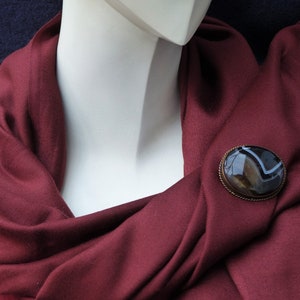 May include: A maroon scarf with a brown and white agate brooch. The brooch is oval shaped and has a silver setting.
