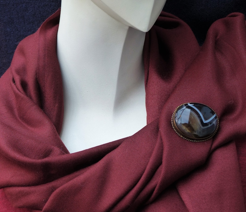 May include: A maroon scarf with a brown and white agate brooch. The brooch is oval shaped and has a silver setting.
