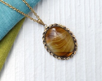 May include: A vintage gold-tone necklace with a large oval pendant. The pendant features a polished, striped stone in shades of brown and amber, set in a decorative, twisted-rope style frame. The necklace chain is also gold-toned.