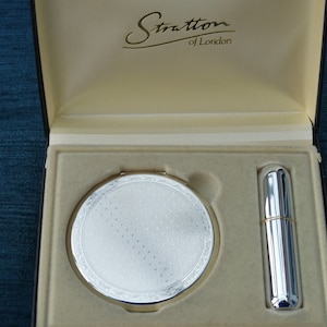 Puede incluir: Un espejo compacto plateado y un tubo de lápiz labial plateado en una caja de color crema con el texto "Stratton of London" en la tapa.