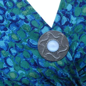 May include: A silver-toned brooch with a blue glass center, featuring a star-shaped design. The brooch is pinned to a blue and green patterned fabric.