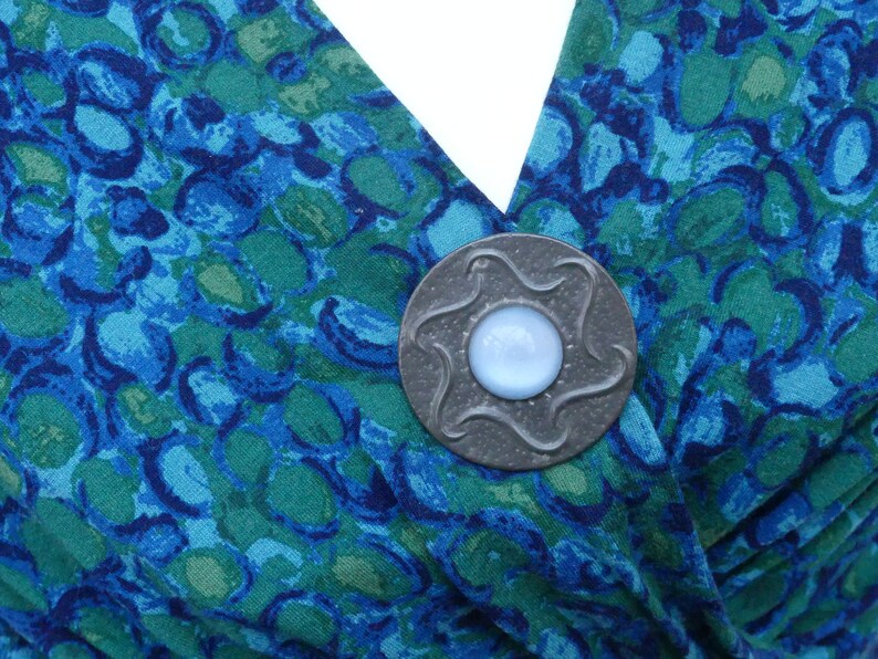 May include: A silver-toned brooch with a blue glass center, featuring a star-shaped design. The brooch is pinned to a blue and green patterned fabric.