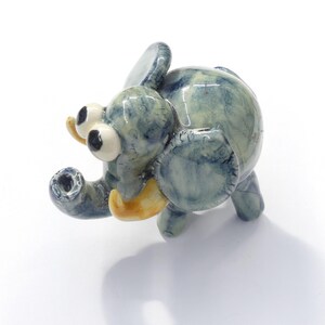 May include: A small ceramic elephant figurine with a blue and green marbled glaze. The elephant has large, white eyes and a yellow trunk.