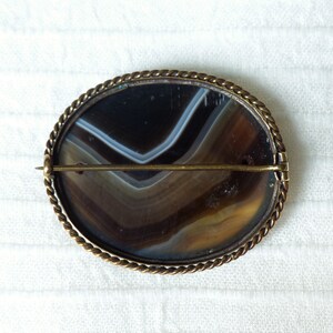 May include: A brown and white agate cameo brooch with a gold-toned metal setting. The brooch has a rope-like border and a pin on the back.