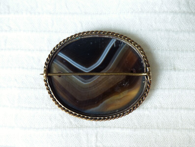 May include: A brown and white agate cameo brooch with a gold-toned metal setting. The brooch has a rope-like border and a pin on the back.