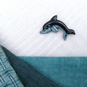 Puede incluir: Una pequeña figura de delfín de cerámica con un esmalte negro y azul. El delfín está saltando fuera del agua con la boca abierta.
