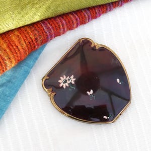 Puede incluir: Un espejo compacto vintage con un marco dorado y una base de esmalte morado oscuro. El compacto está decorado con flores rosas y blancas pintadas a mano.