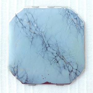 Op de afbeelding: Een zilveren compact met een blauw en wit marmerpatroon.