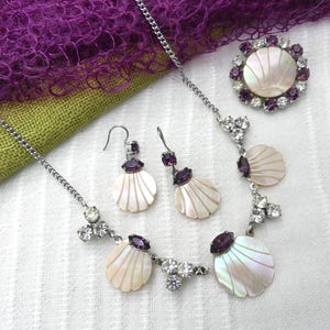 Puede incluir: Un conjunto de joyas plateadas que incluye un collar, pendientes y un broche. El conjunto presenta colgantes en forma de concha y detalles de piedras preciosas moradas y transparentes. Las joyas se exhiben sobre una superficie blanca con un fondo de tela morada y verde.