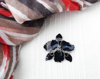 May include: A black orchid brooch with silver detailing. The brooch is on a white fabric background.