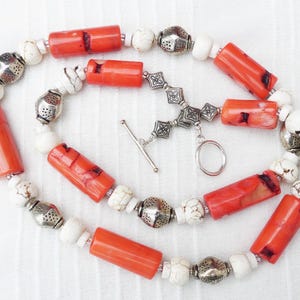 Op de afbeelding: Een lange ketting met oranje en witte cilindrische kralen en zilveren metalen kralen. De ketting heeft een zilveren metalen schuifgesp.