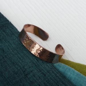 May include: A hammered copper cuff bracelet. The bracelet has a textured surface and a warm, reddish-brown colour. It is displayed on a white surface with a dark teal, teal, and olive green fabric.