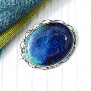 May include: Oval brooch with a silver-tone frame and a large, blue and green stone. The stone has a marbled appearance with shades of blue and green. The brooch is set against a white surface with folded fabric in shades of blue and green.