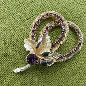 May include: A vintage gold-tone brooch featuring a leaf design and two interlocking circles, each studded with small purple rhinestones. A large purple rhinestone is set at the leaf's base. The brooch is displayed on a green backdrop.