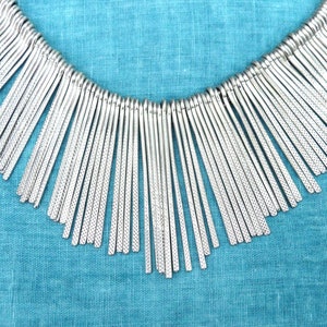 Puede incluir: Un collar plateado con un diseño de flecos. El collar está hecho de muchas hebras de metal pequeñas y delgadas que cuelgan de un punto central. Las hebras son todas de la misma longitud y están espaciadas uniformemente. El collar está sobre un fondo de tela azul.