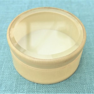 Könnte beinhalten: Eine runde Holzkiste mit einem durchsichtigen Deckel. Die Kiste ist aus hellem Holz gefertigt und hat eine glatte Oberfläche.