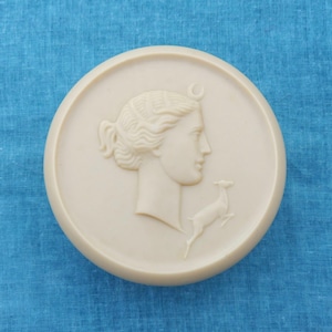 May include: A white ceramic round plaque with a raised relief design of a woman's profile with a crescent moon above her head and a deer below her.