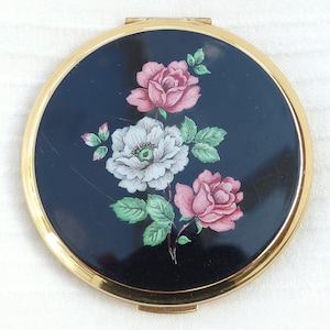 May include: A gold compact mirror with a black background and a floral design featuring pink, white, and green roses.