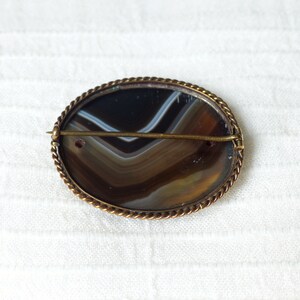 May include: A brown and white agate cameo brooch with a gold-toned metal setting. The brooch has a rope-like border and a pin on the back.