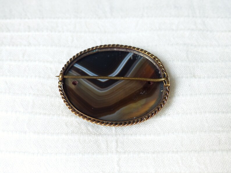 May include: A brown and white agate cameo brooch with a gold-toned metal setting. The brooch has a rope-like border and a pin on the back.