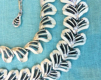 Puede incluir: Un collar y colgante a juego de tono plateado vintage. El collar presenta un diseño repetitivo en forma de hoja con esmalte blanco y detalles en negro. El fondo es una tela azul claro.