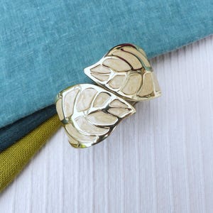 Peut inclure: Deux bracelets de manchette dorés en forme de feuilles avec un remplissage en émail crème. Les bracelets sont posés sur une surface en bois blanc avec un tissu bleu sarcelle, vert foncé et vert olive en arrière-plan.