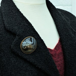 May include: A black blazer with a brown and white agate stone brooch. The brooch is oval shaped and has a gold tone setting.