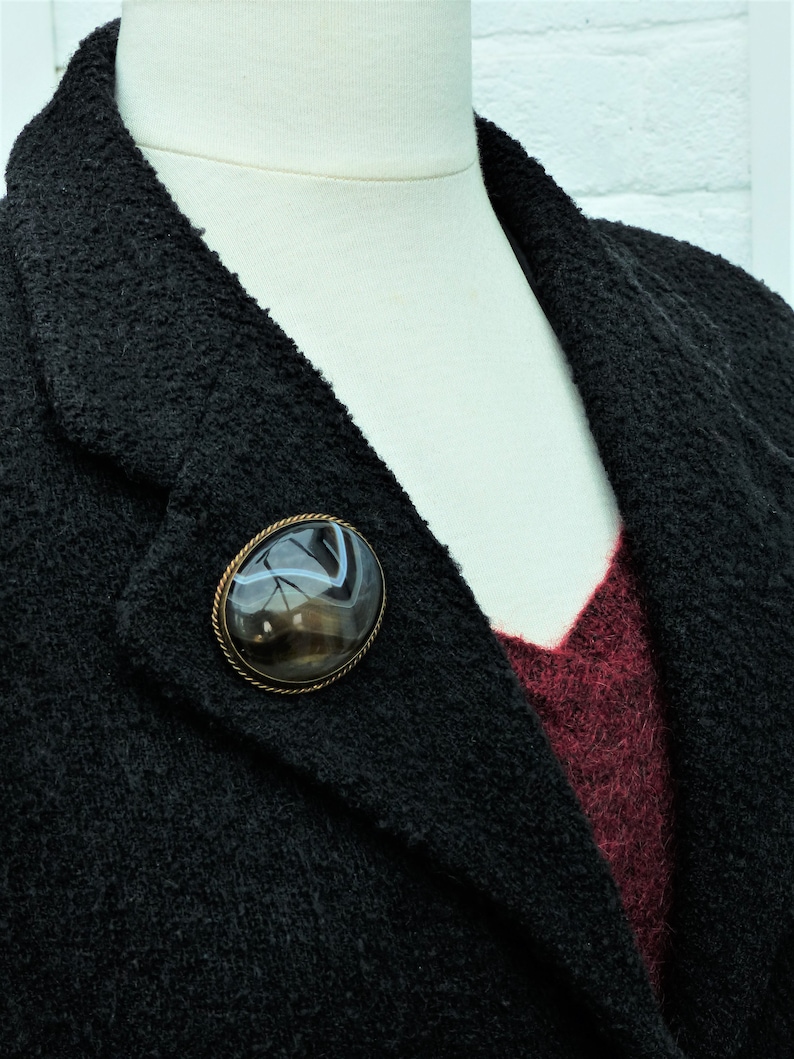 May include: A black blazer with a brown and white agate stone brooch. The brooch is oval shaped and has a gold tone setting.