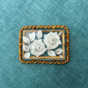 Pode incluir: Broche retangular vintage com borda de corda torcida dourada. O broche apresenta um centro de vidro transparente com detalhes de rosas e folhas brancas. O fundo é um tecido de cor verde-azulado.