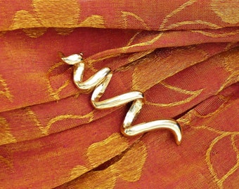 May include: A gold-colored, twisted, metal brooch in the shape of a snake, resting on a red and gold fabric with a floral pattern.