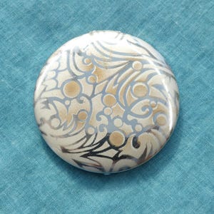 May include: A round, white compact with a silver, swirling, floral design.