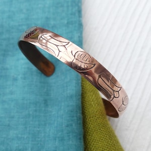 May include: Copper cuff bracelet with an etched floral design. The open-ended bracelet has a polished finish and a detailed pattern of leaves and flowers. The jewellery is displayed on a blue, green, and white background.