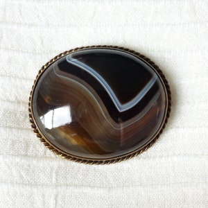 May include: A brown and white agate gemstone brooch with a gold-toned metal setting. The brooch has a rope-like border.