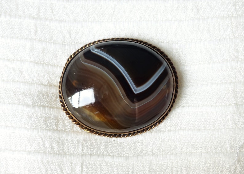 May include: A brown and white agate gemstone brooch with a gold-toned metal setting. The brooch has a rope-like border.