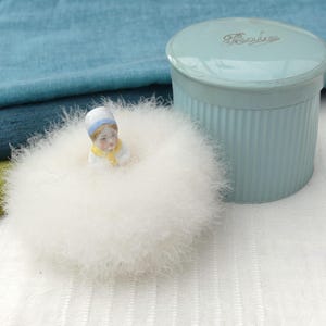 May include: A light blue powder puff container with the word "Modes" printed on the lid. The container is open and a white fluffy powder puff is inside. A small porcelain doll wearing a blue and white sailor outfit is sitting on top of the powder puff.
