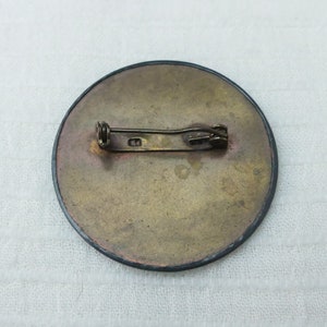 May include: The back of a round, brass-colored pin with a black border. The pin has a simple, straight metal clasp.
