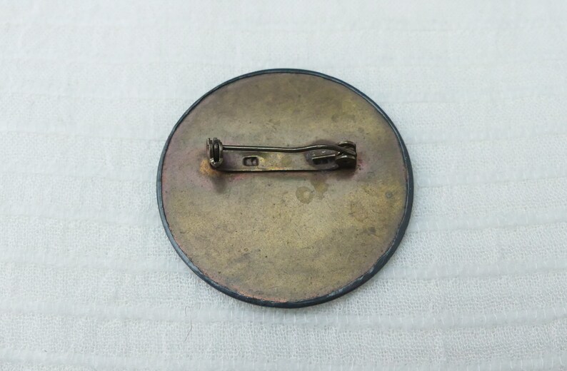 May include: The back of a round, brass-colored pin with a black border. The pin has a simple, straight metal clasp.