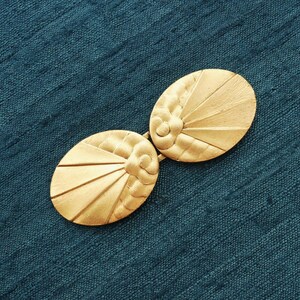 Può includere: Una spilla o chiusura dorata in stile Art Déco. Il pezzo presenta due componenti ovali collegati da una cerniera. Ogni ovale ha un design a forma di ventaglio con un elemento floreale centrale. Lo sfondo è un tessuto strutturato color verde acqua.