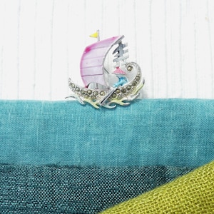 Puede incluir: Broche de barco vikingo plateado con vela rosa y bandera amarilla. El broche presenta detalles decorativos y está colocado sobre telas azules y verdes. Una pequeña figura es visible dentro del barco.