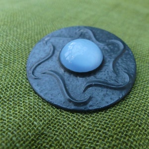 May include: A round, dark gray metal pin with a wavy design. The center of the pin features a light blue, round, glass cabochon.