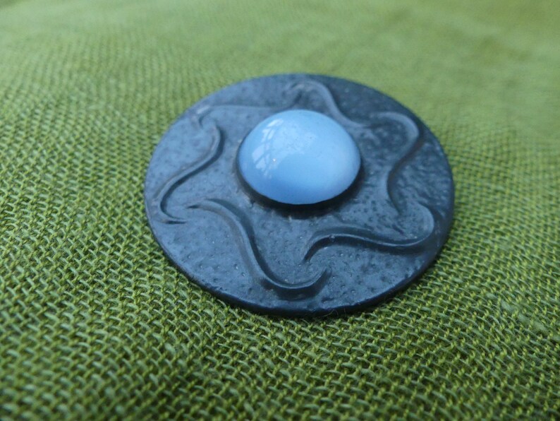 May include: A round, dark gray metal pin with a wavy design. The center of the pin features a light blue, round, glass cabochon.
