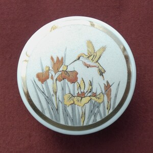 Può includere: Una scatola portaoggetti in ceramica bianca con bordo dorato e un disegno dipinto di un colibrì che vola sopra iris gialli e arancioni.