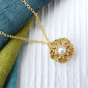 May include: A gold-colored pendant necklace with a round, ornate design and a central pearl. The necklace chain is also gold. The pendant is set against a backdrop of blue and green fabric and white fabric.