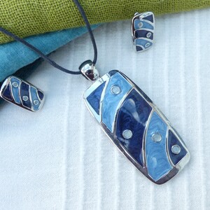 Puede incluir: Un collar de plata con un colgante de esmalte azul y blanco y pendientes a juego. El colgante es rectangular y presenta un diseño de remolino con puntos blancos.