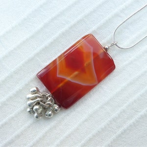 May include: A silver chain necklace with a rectangular carnelian pendant and a silver tassel charm. The carnelian is red, orange, and white.