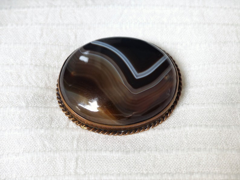 May include: A brown agate gemstone brooch with a copper-toned metal setting. The agate has a swirling pattern of brown, black, and white.