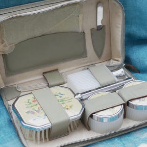 Könnte beinhalten: Eine graue Leder-Reise-Kosmetiktasche mit Reißverschluss. Die Tasche ist geöffnet und enthält verschiedene Toilettenartikel, darunter eine Bürste, einen Kamm, eine Flasche Lotion und zwei kleine Gläser. Die Tasche ist mit einem hellgrauen Stoff ausgekleidet.