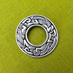 May include: A silver-coloured, circular pendant with an intricate, embossed design. The pendant features a central hole and is set against a textured, olive-green background. The design appears to be of Celtic or Viking origin.